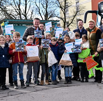 Eine Gruppe von Volksschülern sowie acht Erwachsene schauen lachend in die Kamera. Alle halten ein Buch mit dem Titel 