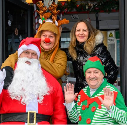 Weihnachtsmann, Rentier, Weihnachtswichtel und Projektleiterin des KMG-Weihnachtsbusses lächeln frontal in die Kamera