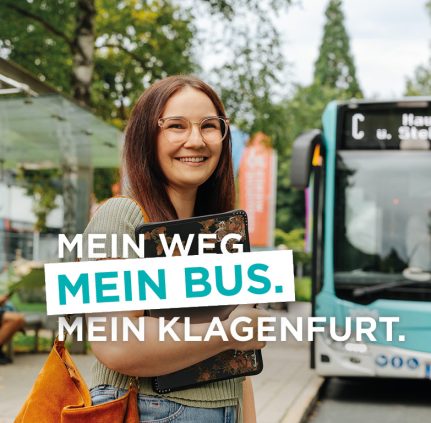 Hier sieht man ein fertiges Sujet der neuen KMG-Personenkampagne. Eine junge Frau mit dunklen langen Haaren und Brille hält ein iPad im Arm und lächelt in die Kamera. Etwas entfernt im Hintergrund sieht man eine Bushaltestelle und einen KMG-Bus. Der Kampagnentitel lautet: Mein Weg. Mein Bus. Mein Klagenfurt.