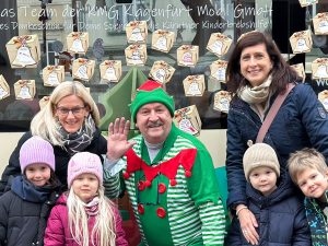 Vier Kindergartenkinder stehen vor dem KMG-Weihnachtsbus. In der Mitte steht ein Buslenker, der als Wichtel verkleidet ist. Hinter den Kindern stehen zwei Damen. Alle lächeln in die Kamera, der Wichtel winkt. Hinter den Personen sieht man einen Teil der Fensterscheibe des Busses, auf dem zahlreiche Weihnachtspaket-Aufkleber zu sehen sind, die die Leute aufgeklebt haben.