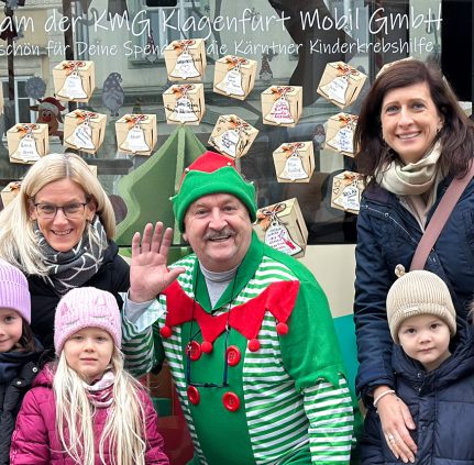 Vier Kindergartenkinder stehen vor dem KMG-Weihnachtsbus. In der Mitte steht ein Buslenker, der als Wichtel verkleidet ist. Hinter den Kindern stehen zwei Damen. Alle lächeln in die Kamera, der Wichtel winkt. Hinter den Personen sieht man einen Teil der Fensterscheibe des Busses, auf dem zahlreiche Weihnachtspaket-Aufkleber zu sehen sind, die die Leute aufgeklebt haben.