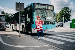 Eine junge, blonde Frau steht lächelnd vor einem KMG-Bus. Sie trägt ein Fan-T-Shirt des EC-KAC sowie einen Fan-Schal und eine Fan-Haube, in der Hand hält sie 2 KAC-Eintrittskarten.