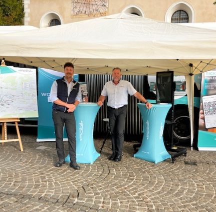 Zwei Männer stehen unter einem beigen Zeltpavillon, angelehnt an 2 türkise Stehtische und lächeln in die Kamera. Dahinter sieht man Teile eines Busses, eine große Schautafel mit dem KMG-Liniennetz sowie ein Roll Up mit dem Sujet der KlagenfurtMobil 2.0-App.