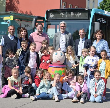 Eine Schulklasse mit 19 Kindern und 6 Erwachsenen vor einem KMG-Bus. Die 8 Kinder in der ersten Reihe sitzen am Boden, die anderen Kinder stehen dahinter in der zweiten Reihe. In der dritten Reihe stehen Erwachsenen. Im Hintergrund sieht man die Front eines KMG-Busses und daneben eine Haltestelle.