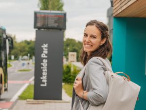 KMG-Mobilitätsknoten Lakeside Park, links fährt ein Bus an der Haltestelle vorbei an der eine junge Dame mit Rucksack lächelnd in die Kamera schaut.