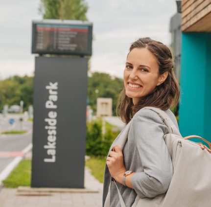 KMG-Mobilitätsknoten Lakeside Park, links fährt ein Bus an der Haltestelle vorbei an der eine junge Dame mit Rucksack lächelnd in die Kamera schaut.