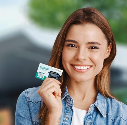 Junge Frau lacht in die Kamera und hält dabei eine KMG-Prepaid Card in der rechten Hand. Im Hintergrund sieht man verschwommen das Dach einer Bushaltestelle sowie die Blätter von Bäumen.