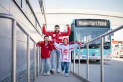 Eine junger Mann und vor ihm zwei Buben im Alter von ca. 8 Jahren gehen lachend eine Rampe hoch. Daneben steht ein KMG-Bus. Der Mann und der Junge links vor ihm tragen eine rote Jacke mit KAC-Logo sowie einen Fan-Schal. Der Junge rechts vorne trägt einen Fan-Schal und ein weiß/rotes T-Shirt des EC-KAC.